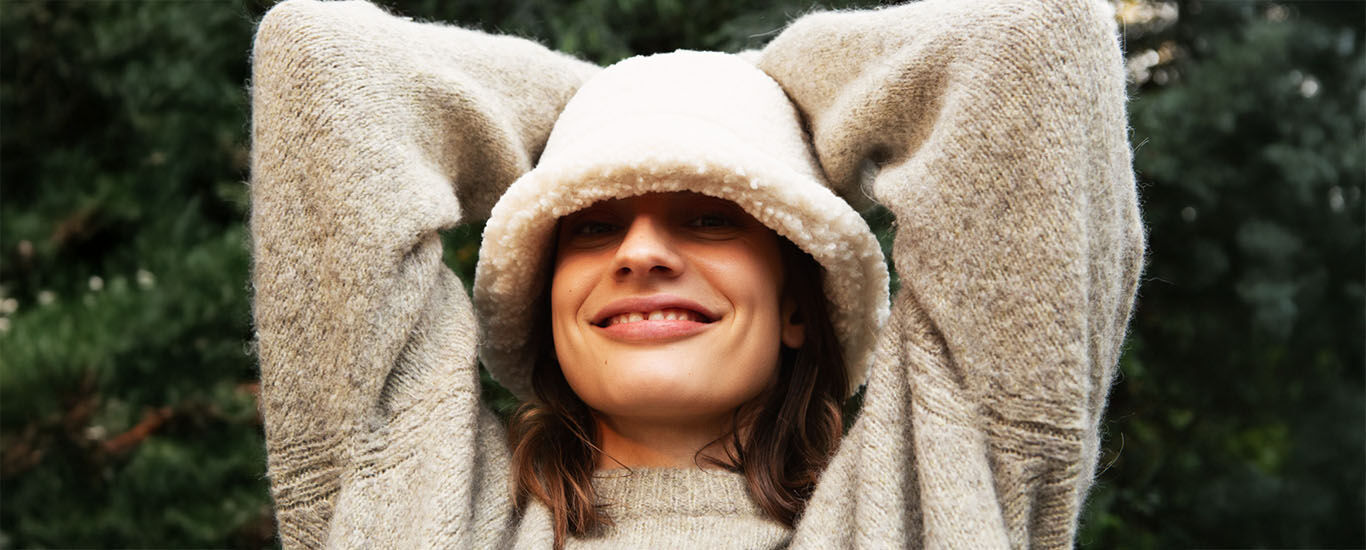 Woman in cozy sweater with hood smiles relaxed and stretches her arms over her head.